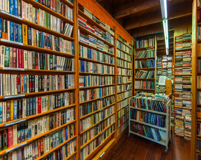 The image shows a narrow aisle flanked by tall bookshelves filled with numerous books, with a cart holding additional books in the center.