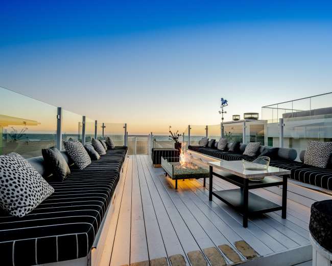A rooftop terrace features a long seating area with striped cushions, a central coffee table, and a fire pit, all set against a sunset view over the ocean.