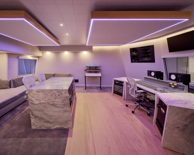 The image shows a modern music recording studio featuring a sleek control desk, comfortable seating area, and ambient lighting.