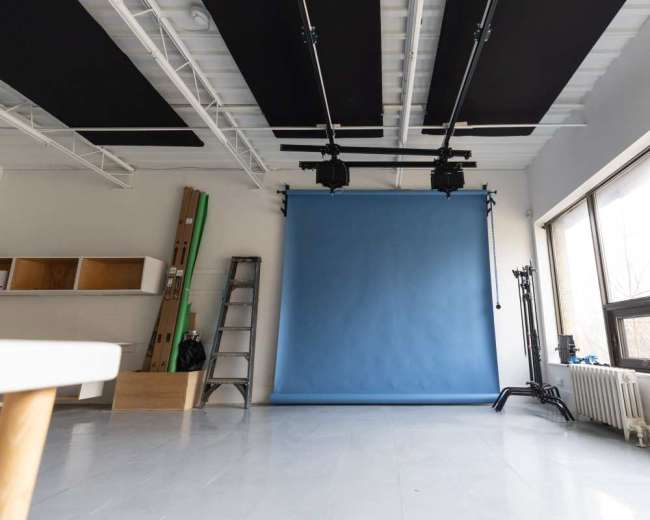 The image shows a minimalist photography studio featuring a blue backdrop, a ladder, lighting equipment, and large windows.