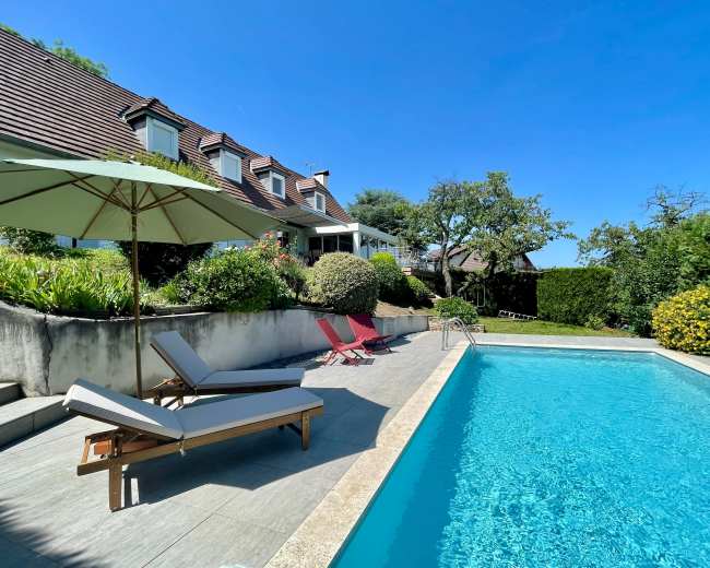 The image shows a swimming pool bordered by lounge chairs and an umbrella in a backyard garden with a house and trees in the background.