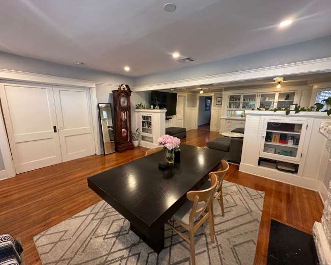 The image shows a well-lit room featuring a dining table with chairs, a decorative clock, a television, and built-in shelves against the walls.