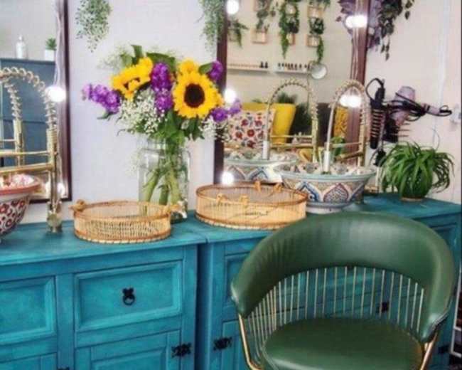 The image shows a turquoise cabinet with a green chair and a vase of sunflowers on a countertop, surrounded by mirrors and decorative items.