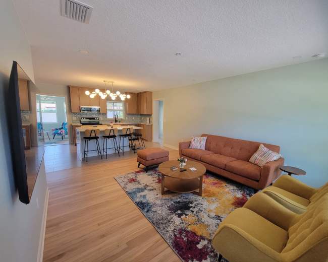 The image shows a modern, open-concept living area with a couch, two armchairs, a coffee table, and a kitchen visible in the background.