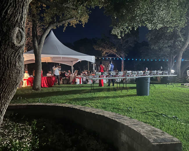 A tent is set up in a garden at night, with guests seated at tables and a buffet spread laid out in the foreground.