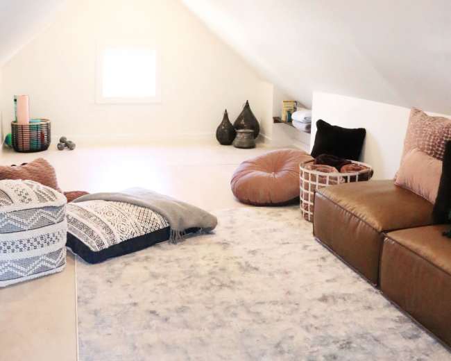 The image shows a cozy attic space with a slanted ceiling, featuring scattered floor cushions, a small couch, and decorative items arranged on shelves.