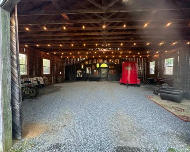 The image shows the interior of a spacious barn-like structure with exposed beams, gravel flooring, and string lights, featuring decor items along the walls and a red trailer parked inside.