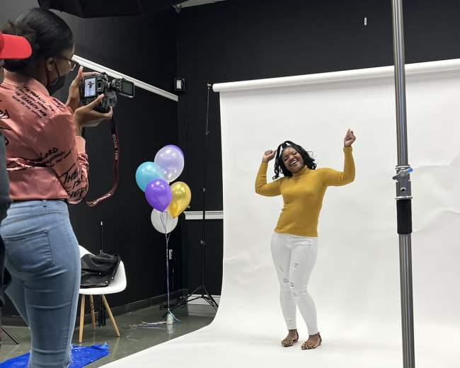 A person wearing a yellow sweater poses playfully in front of a white backdrop while another individual captures the moment with a camera.