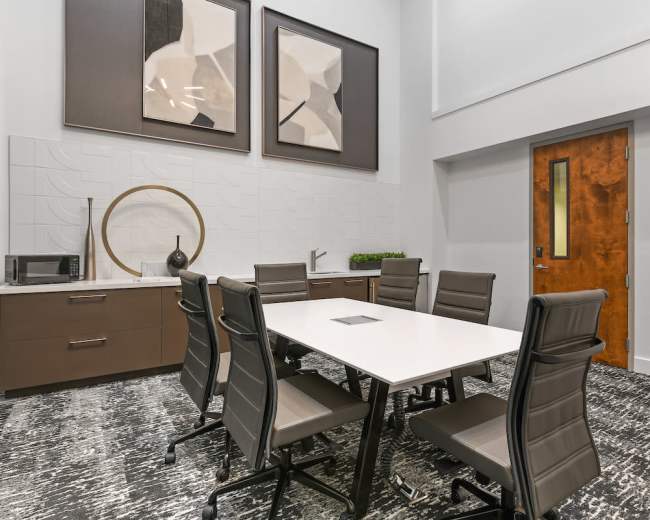 A modern conference room features a long white table surrounded by six leather chairs, with abstract art on the walls and appliances in a cabinet.