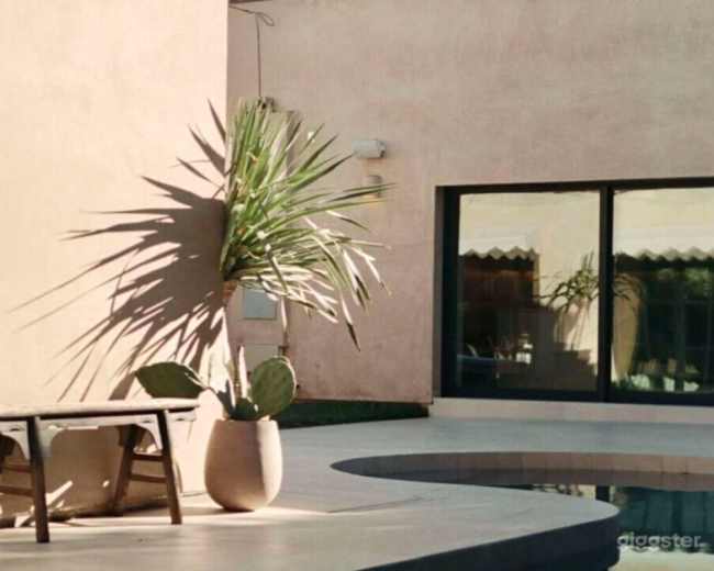 The scene features a minimalist outdoor space with a small wooden table beside a large potted plant and a swimming pool, set against a smooth, beige wall.