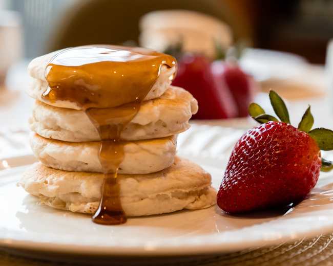 A stack of four pancakes topped with syrup and a fresh strawberry sits on a white plate.