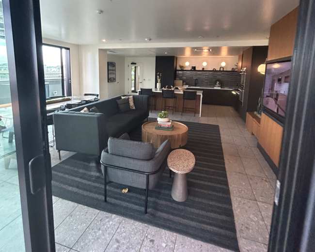 The image shows a modern, open-concept living area with a kitchen, featuring a gray couch, a round wooden coffee table, and large windows.