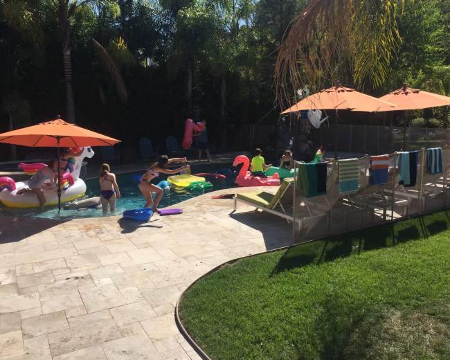 A group of children enjoys playing in a backyard pool surrounded by colorful floaties, lounge chairs, and sun umbrellas.