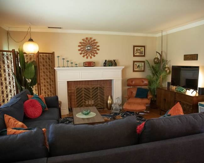 A cozy living room features a sectional sofa, a fireplace with decorative accents, a wooden media console, and various greenery throughout the space.