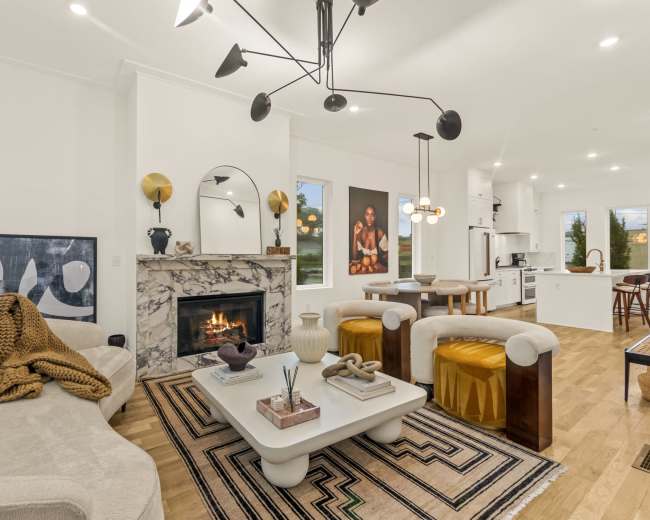 The image shows a modern living room with a marble fireplace, a circular mirror, a beige sofa, and a central coffee table surrounded by yellow accent chairs and a patterned rug.