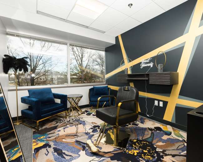 The image shows a modern lounge area with two blue velvet chairs, a round glass table, and a black office chair, set against a geometric gold-accented wall and a large window.