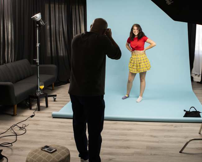 A person is taking a photograph of a model posing in front of a light blue background in a studio.