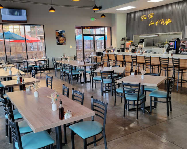 A spacious restaurant interior features rows of tables and chairs, a counter with a menu display, and large windows overlooking an outdoor dining area with umbrellas.