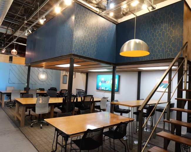 The image shows a modern, open office space with multiple tables, chairs, and a staircase leading to an upper level that features a large screen.
