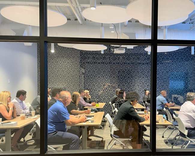 A group of people is seated in a conference room, facing a presenter at the front, with visual aids displayed on a screen.