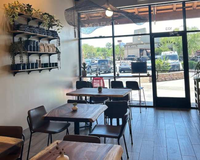 A modern cafe interior with wooden tables, chairs, and plants arranged on a shelf, featuring large glass doors that open to a view of cars outside.