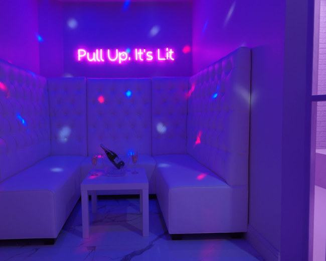 A white seating area with tufted upholstery features a small table and illuminated signage that says "Pull Up. It's Lit," against a purple backdrop.