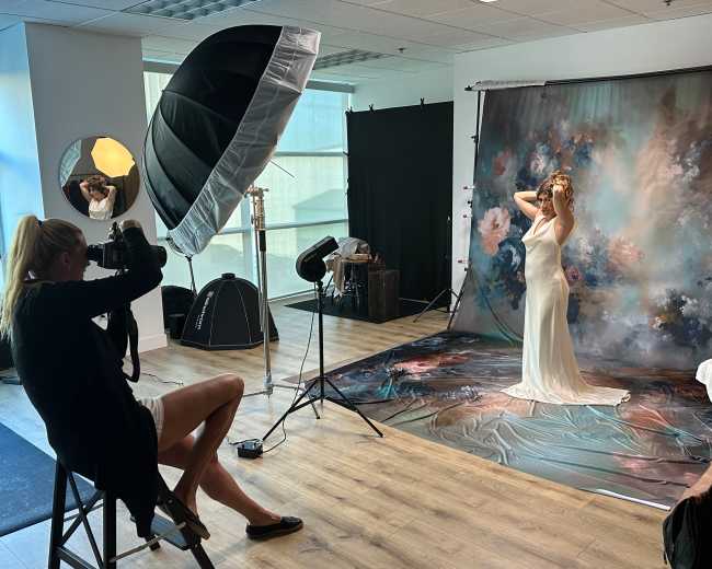 A photographer captures a model in a flowing dress posed in front of a painted backdrop in a studio setting.