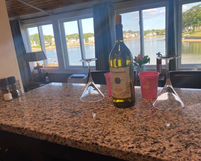 A bottle of wine sits between two wine glasses on a granite countertop overlooking a waterfront view through large windows.
