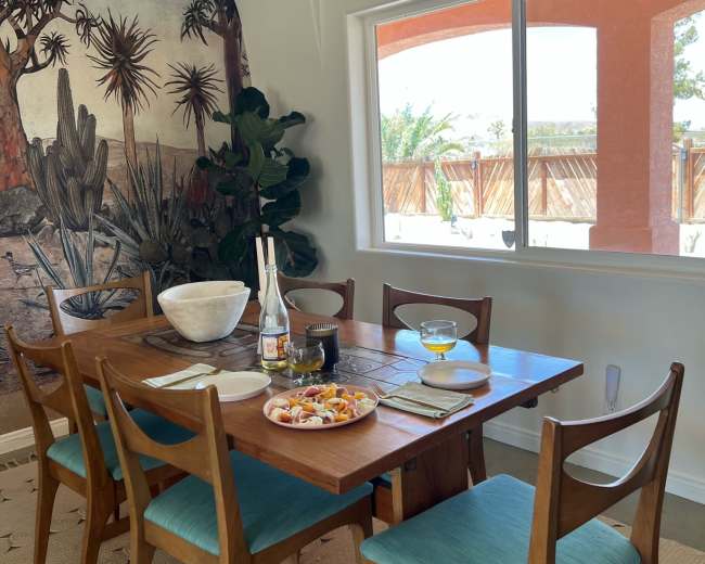 A dining table set with dishes and drinks is positioned near a large window, showcasing a mural of a desert landscape and a green plant in the corner.