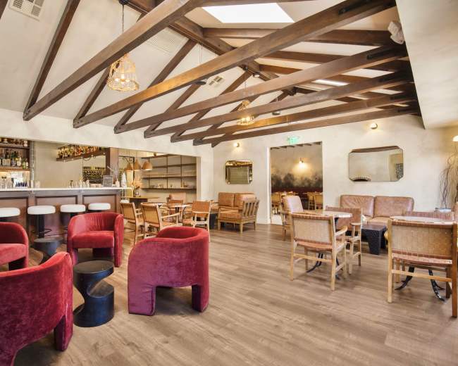 The image shows a modern dining space featuring a bar, seating areas with red chairs, wooden beams, and a warm ambiance.