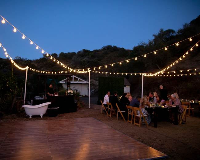 A dinner party is set up outdoors with string lights illuminating the area, featuring a long table and a black serving station, against a backdrop of hills and trees.