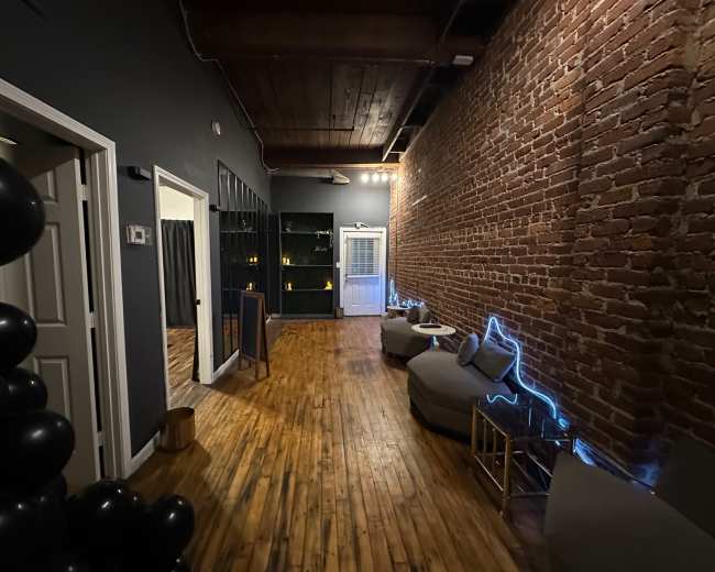 The image shows a dimly lit corridor with exposed brick walls, wooden flooring, and seating areas featuring gray cushions along one side.