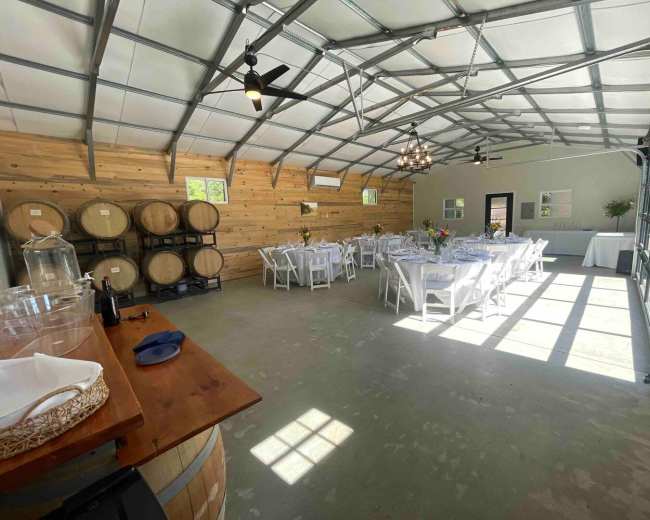 An indoor event space with white tables set for dining, wooden barrels stacked against a wall, and large windows allowing natural light to enter.