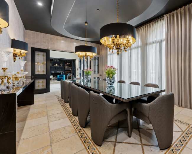 The image shows a modern dining room featuring a long glass table surrounded by upholstered chairs, with large black pendant lights and floor-to-ceiling curtains.