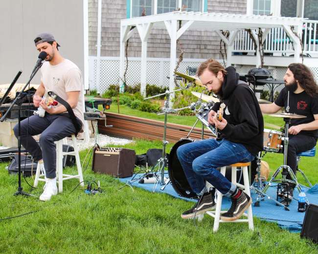 Three musicians are performing outside on a grassy area, with one playing guitar and singing, while the others play bass and drums.
