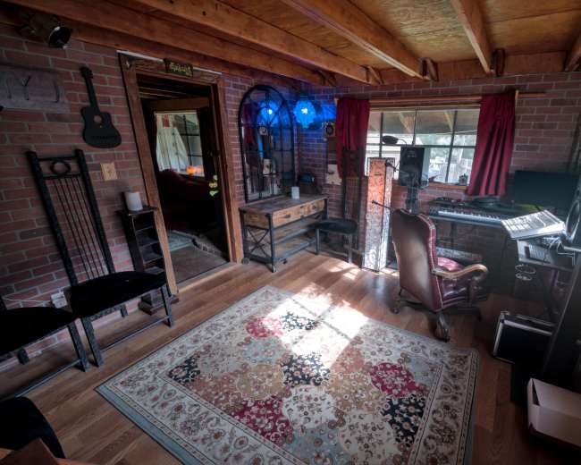 The image depicts a cozy studio workspace with brick walls, a wooden floor, and various musical instruments and equipment arranged around a vibrant area rug.
