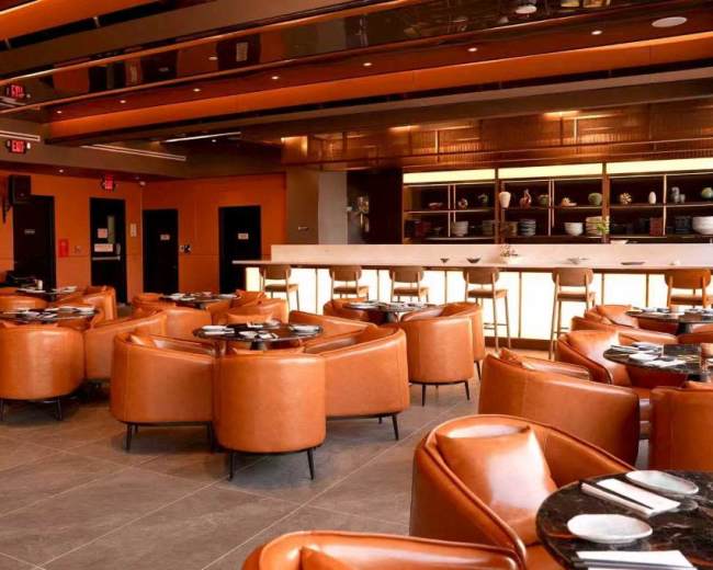 The image shows a modern restaurant interior featuring brown leather seating, a bar with high stools, and a well-lit open area.