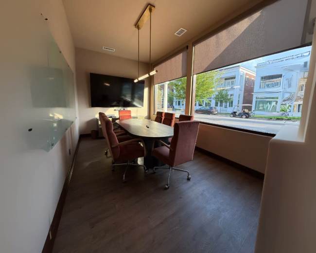 A conference room features a long table surrounded by chairs, large windows displaying a street view, and a mounted TV on the wall.