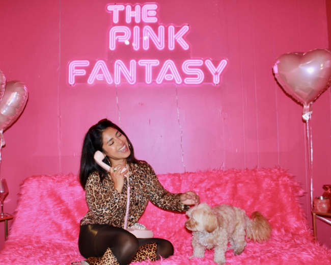 A woman in a leopard-print outfit is sitting on a pink couch talking on a retro telephone while a dog sits beside her, with pink balloons and a sign reading "THE PINK FANTASY" in the background.