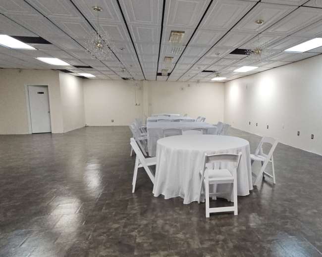 The image shows an empty function room with multiple round tables covered in white tablecloths and white chairs arranged around them.