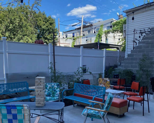 The image shows a backyard seating area featuring various colorful chairs and tables, with string lights overhead and a white fence in the background.