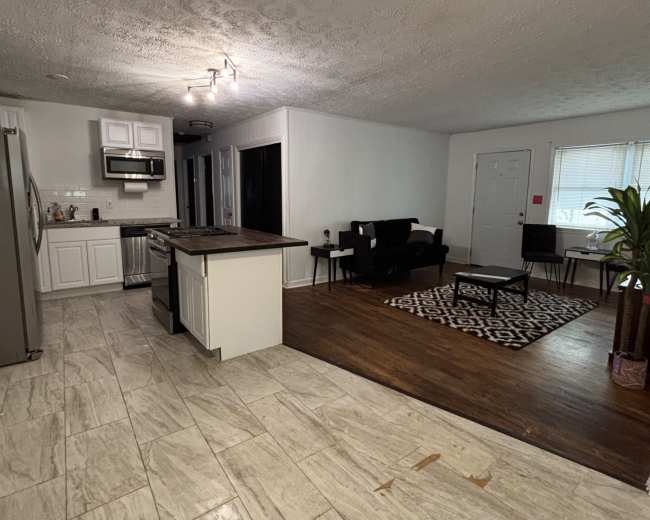 The image shows a spacious living area with a kitchen on one side, featuring tiled floors, a wooden accent area, and various seating arrangements.