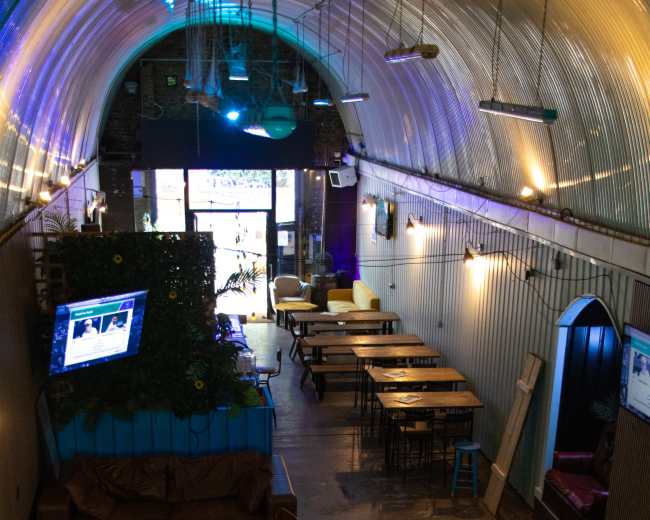 The image shows an interior space of a bar or restaurant featuring wooden tables, a colorful area at the entrance, and multiple screens displaying content on the walls.
