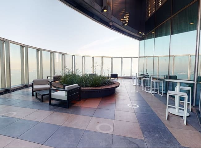 The image shows a modern rooftop terrace with seating and a circular planter, overlooking a scenic view.