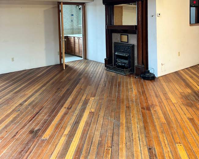 The image shows a spacious, empty room with wooden floors, a fireplace embedded in a wall, and an open doorway leading to a kitchen area.