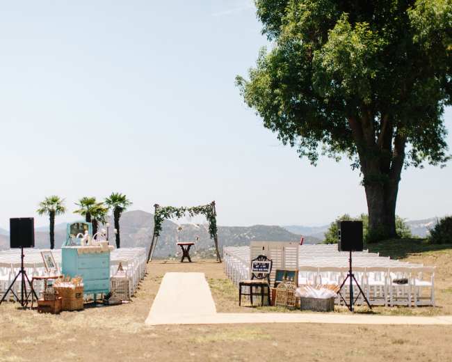 An outdoor wedding ceremony setup features white chairs arranged in rows along a central aisle leading to an arch adorned with greenery and flowers, set against a mountainous backdrop.