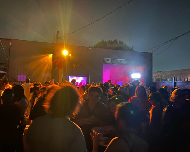 A crowd gathers in an outdoor space with colorful lights and a projection in the background at night.