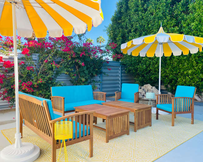 A patio area features bright blue furniture, yellow and white striped umbrellas, and vibrant bougainvillea in the background.
