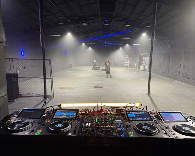 The image shows a DJ setup with controllers and mixers in the foreground, facing an empty warehouse space illuminated by soft lighting and smoke effects.