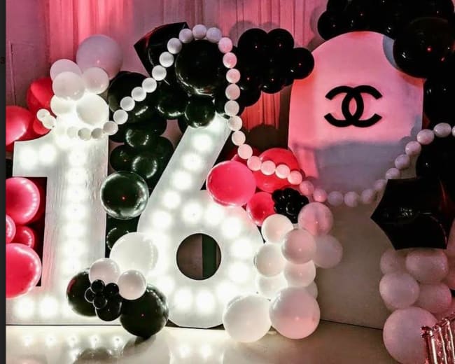 A large, illuminated "16" made of balloons, surrounded by black and white balloons, and displays a Chanel logo in the background.